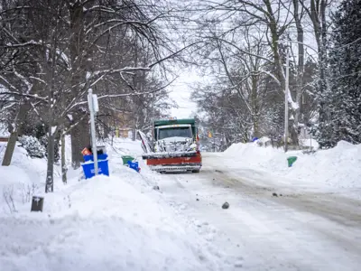 A snowplow travels down a residential street with green bins and recycling set out in the winter