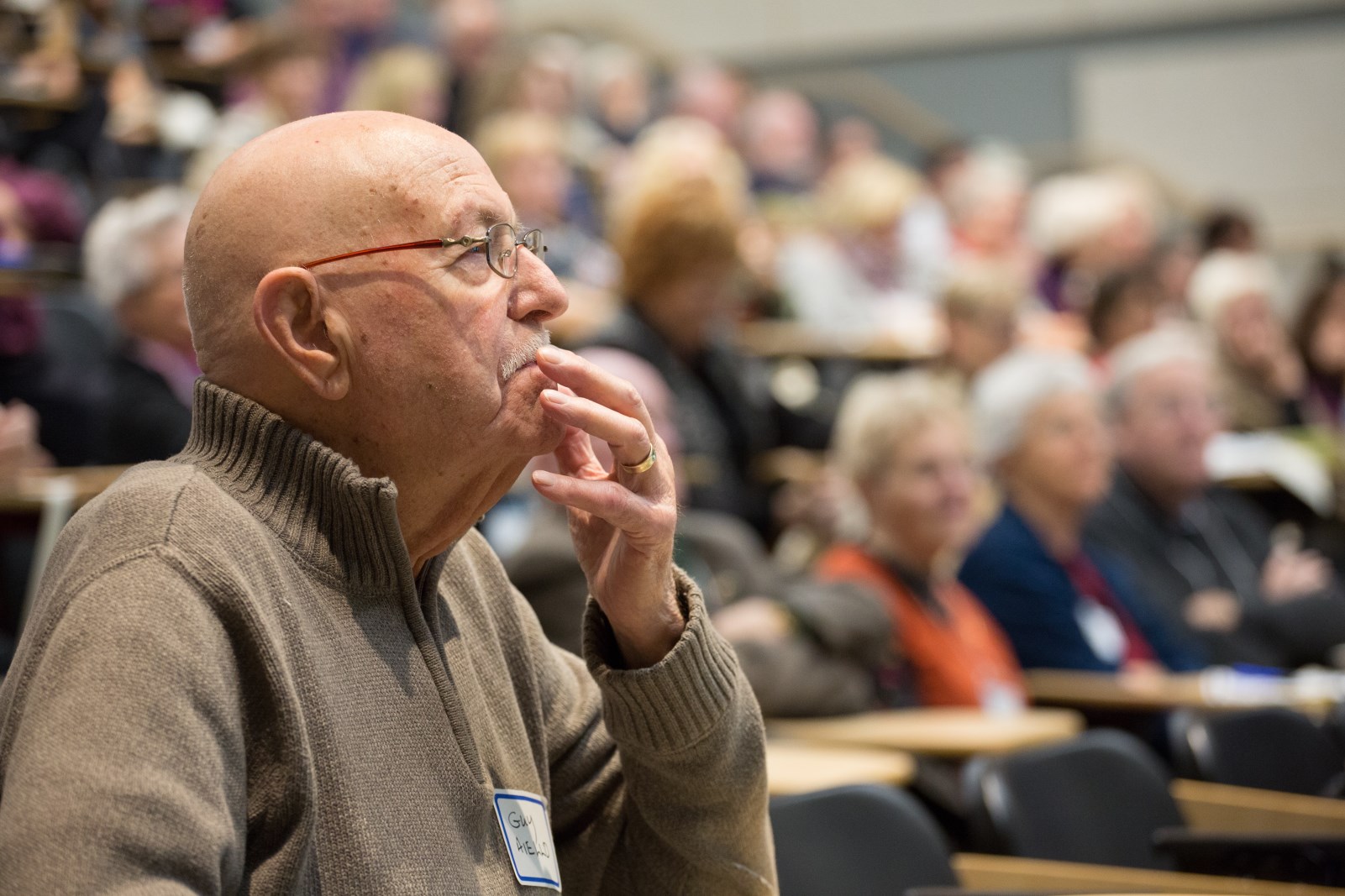 Man sitting in an audience, looking to the right of the image, with his hand at his face, fingers on lips