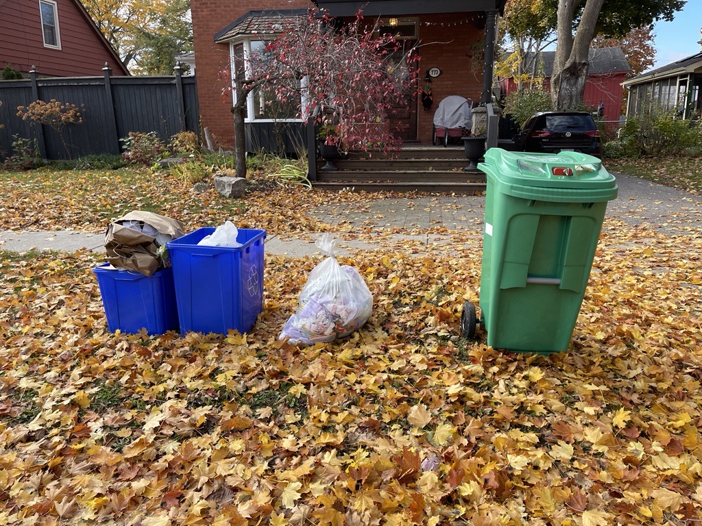 Recycling, clear garbage bag and green bin set out at curb for collection on autumn day