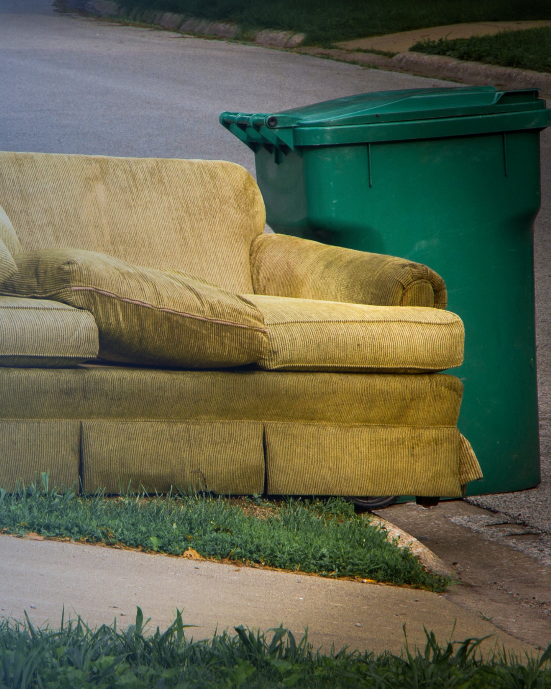 An old couch at side of road next to a Green Bin